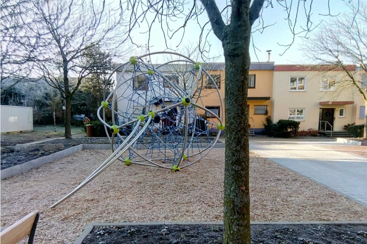 Baum und freier Platz mit Kletter-Kugel aus Stäben und Seilen. Im Hintergrund Reihenhäuser