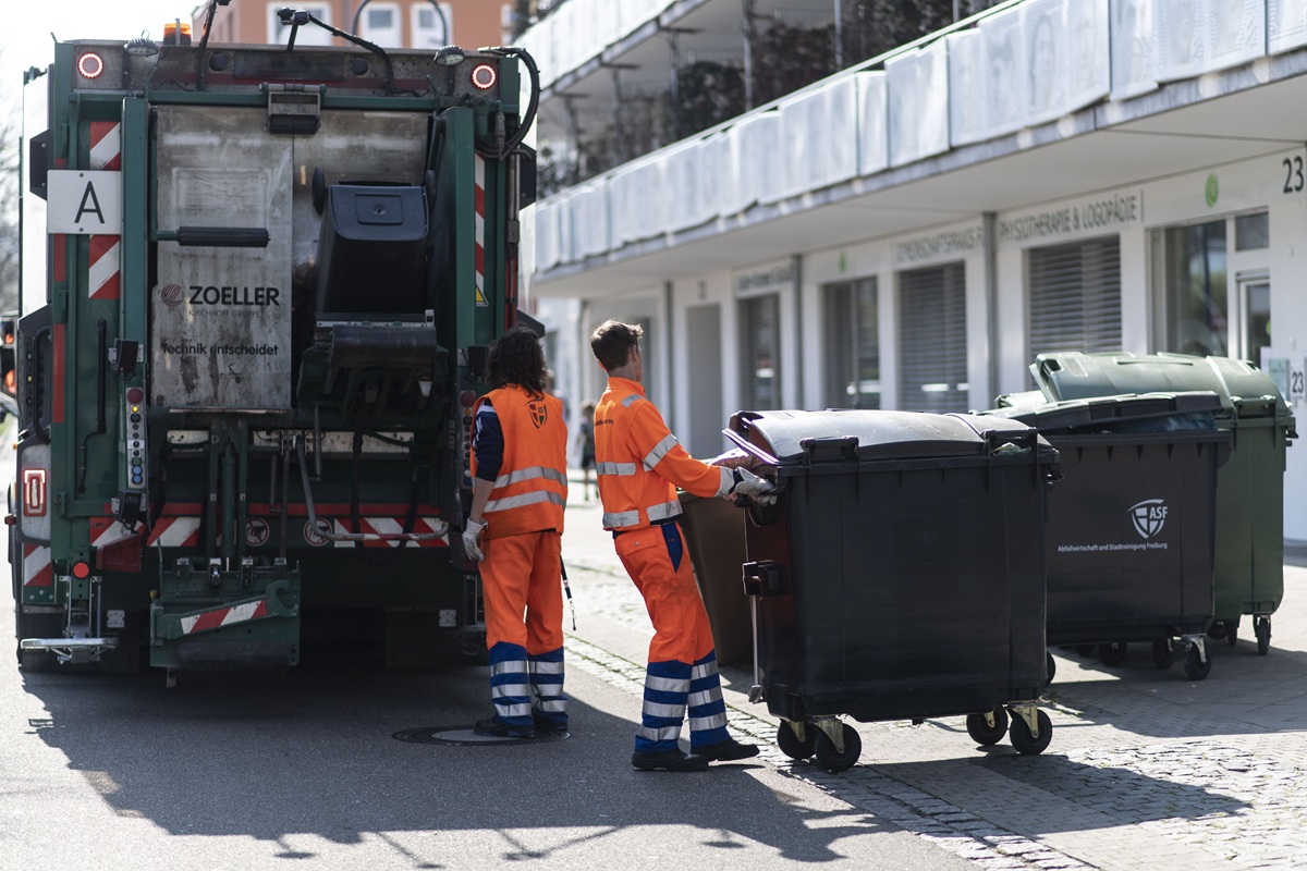 Zwei Müllmänner stehen vor einem Müllauto und laden gerade einen schwarzen Müllcontainer auf.