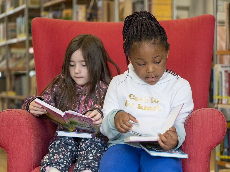 Zwei Mädchen sitzen auf einem roten Sessel und schauen Bücher an.