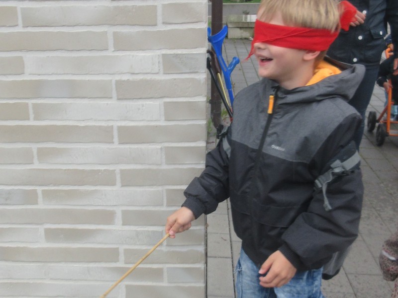 Ein Junge mit verbundenen Augen und einem Stock läuft an einer Häuserecke vorbei.