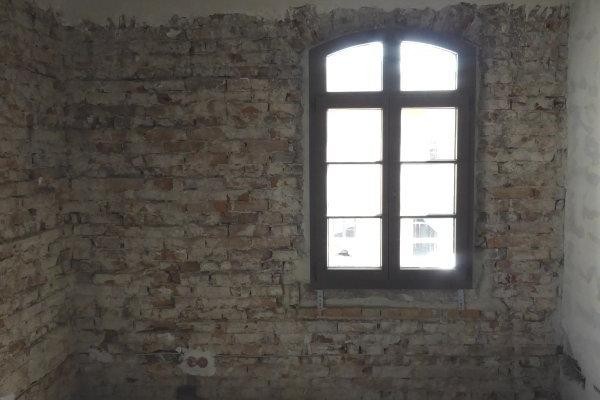 Ieerer Innenraum mit sichtbarer Mauerung durch abgeschlagenem Putz, neues Fenster und rohe Holzbalkendecke