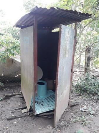 Ein einfaches Toilettenhaus ringsherum Bäume und erdiger Boden