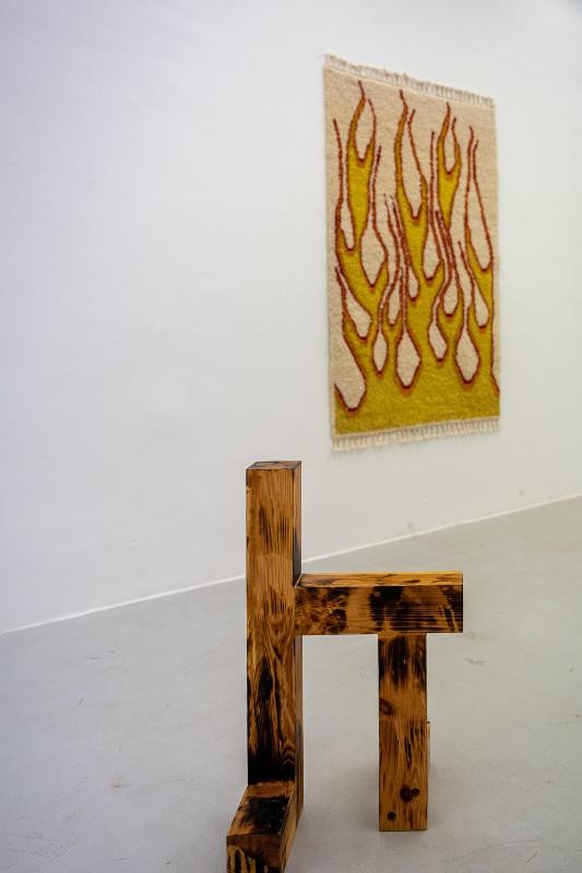 Ausstellungsansicht mit Kunstwerken von Oliver-Selim Boualam (Teppich an der Wann mit Feuermotiv und Holzstuhl)