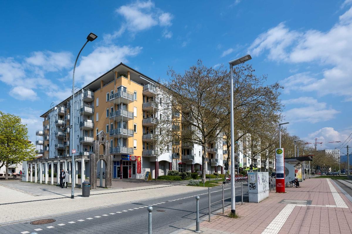 Sanierungsgebiet Betzenhausen-Bischofslinde Straßenbahnhaltestelle, Staße, Straßenbäume, freie Platzfläche mit einer Holz-Tor-Skkulptur und einem Wohn-Eckhausaus
