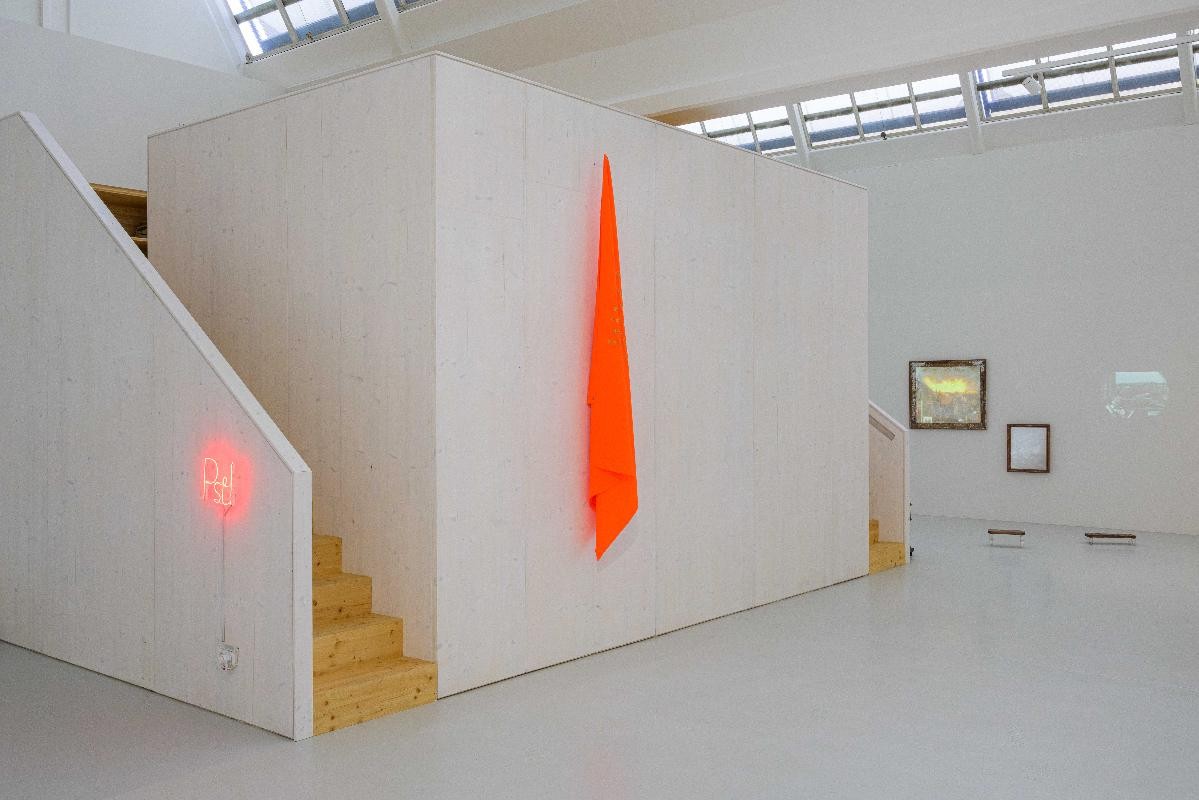 Ausstellungsansicht von "PST!". Es zeigt den Blick in Richtung Eingang. Zusehen ist ein Neonleuchtschild in rot, sowie eine signalrote Fahne und eine Videoinstallation im Hintergrung