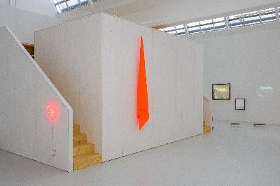 Ausstellungsansicht von "PST!". Es zeigt den Blick in Richtung Eingang. Zusehen ist ein Neonleuchtschild in rot, sowie eine signalrote Fahne und eine Videoinstallation im Hintergrung