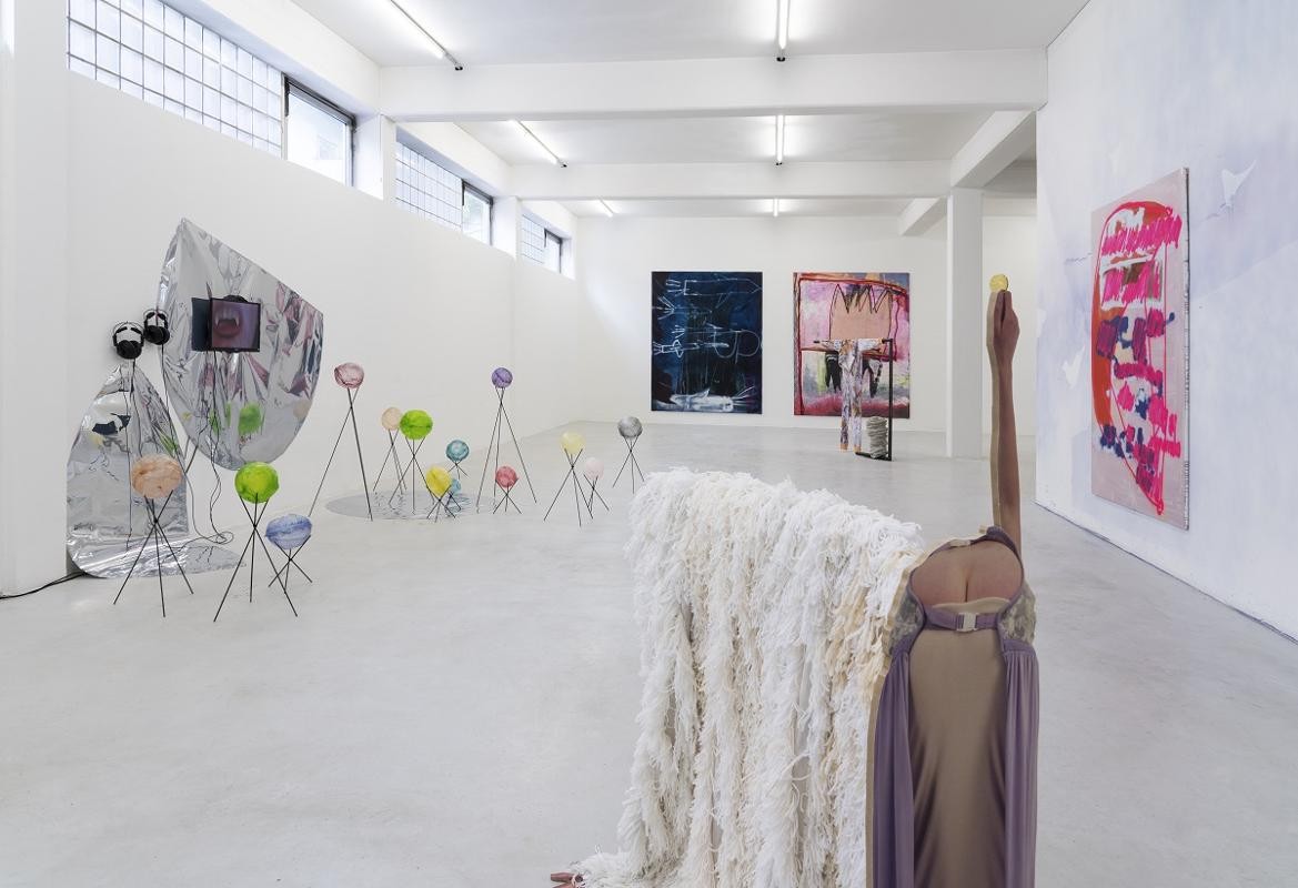  Ausstellungsansicht: im Vorderrund Skulptur mit Flokati und sich bückende Person von hinten, links Videoinstallation mit Blasen aus Reispapier, hinten zwei Leinwände dunkelblau und rosa