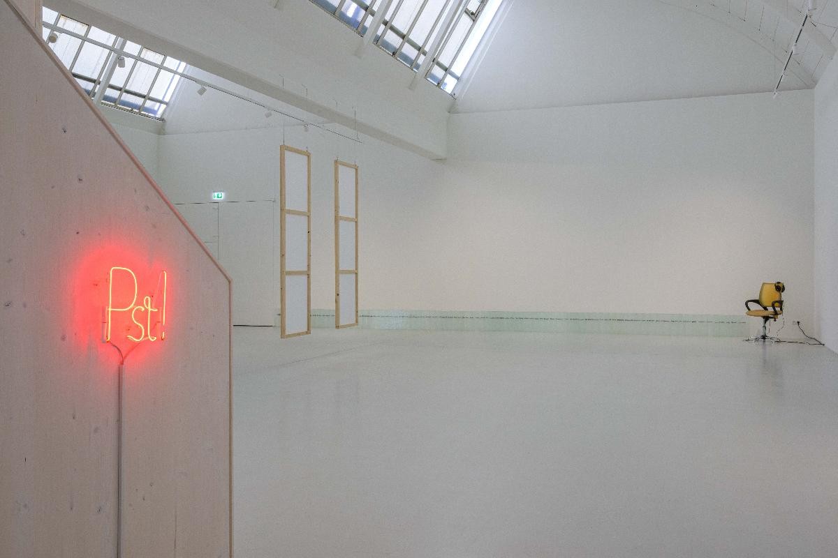 Ausstellungsansicht von "PST!". Es zeigt den Blick in den Raum. Zusehen ist ein Neonleuchtschild in rot vorn und hinten eine schwebende Leinwand sowie am Boden  an der Wand entlang geschichtete Glasplatten