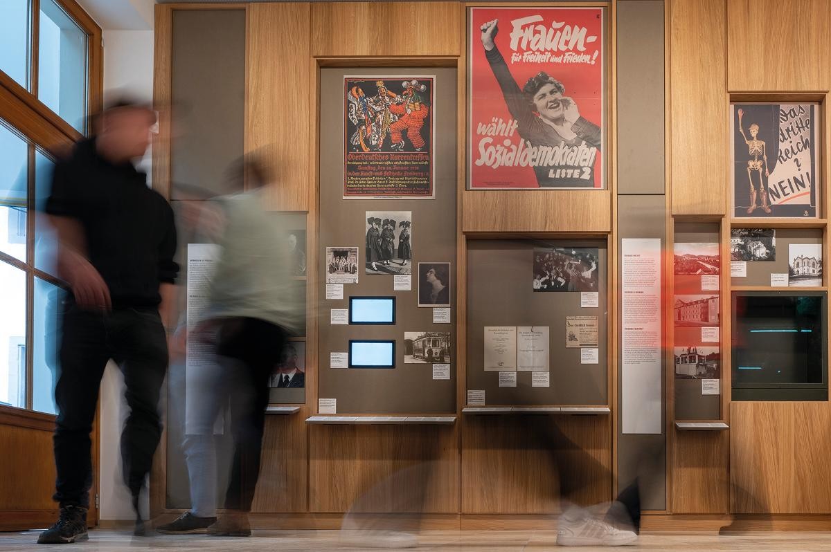 Besucher*innen laufen durch die Ausstellung, im Hintergrund die Ausstellungs-Objekte