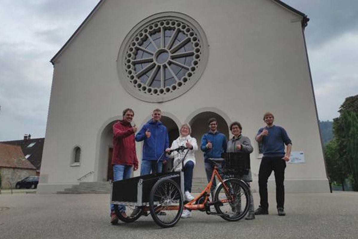 Sechs Menschen, die den Daumen hoch zeigen, vor ihnen ein Lastenrad und hinter ihnen die Dreifaltigkeitskirche.