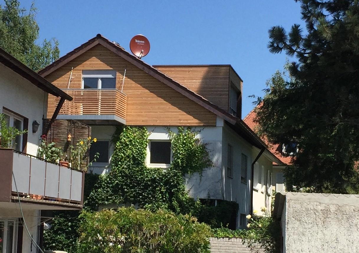 Man sieht eine saniertes Gebäude mit Holzfassade im obersten Stockwerk, einen neuen Balkon und einer Gaube