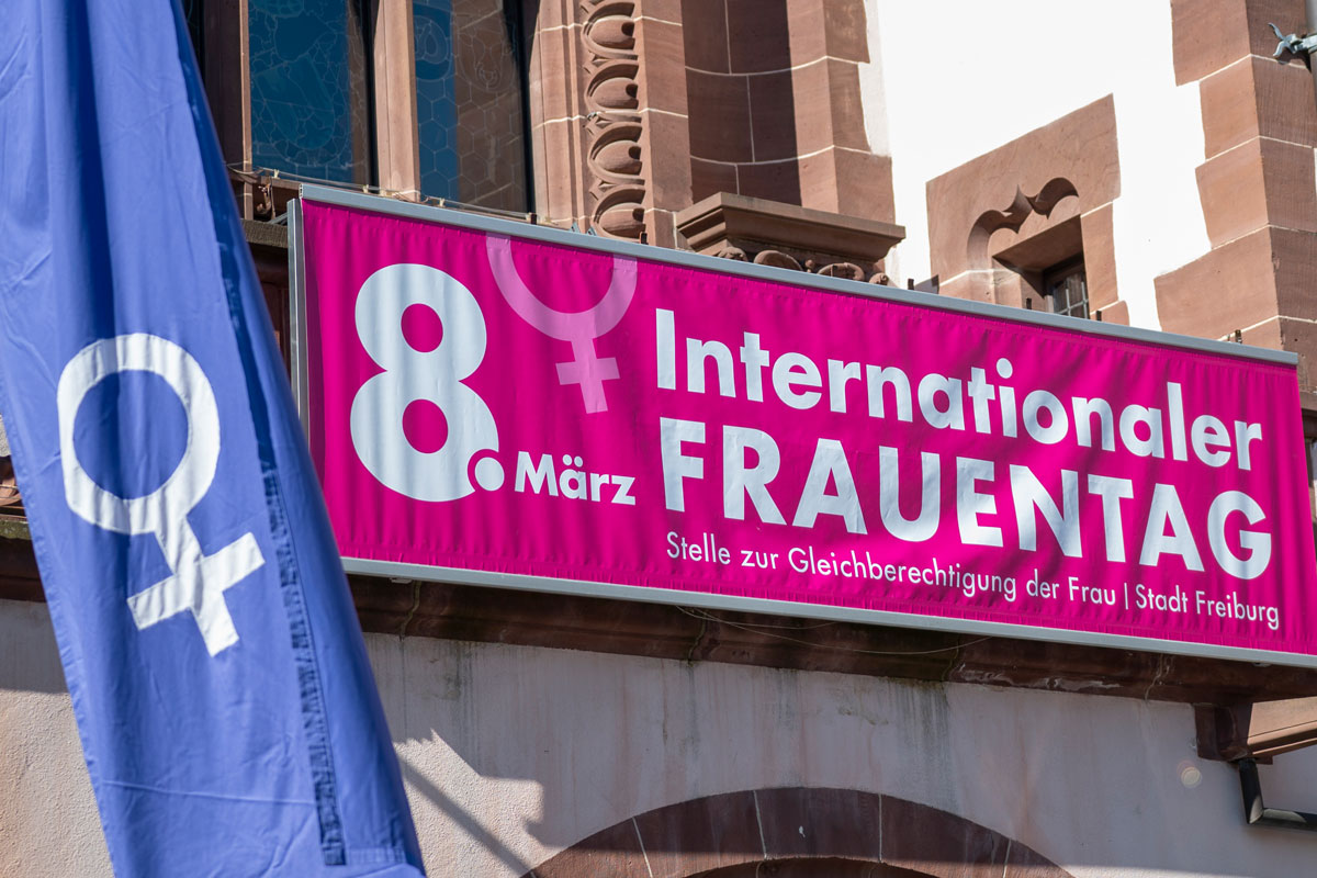Balkon mit pinkem Banner: 8. März, Internationaler Frauentag; blaue Fahne mit Weiblichkeitssymbol