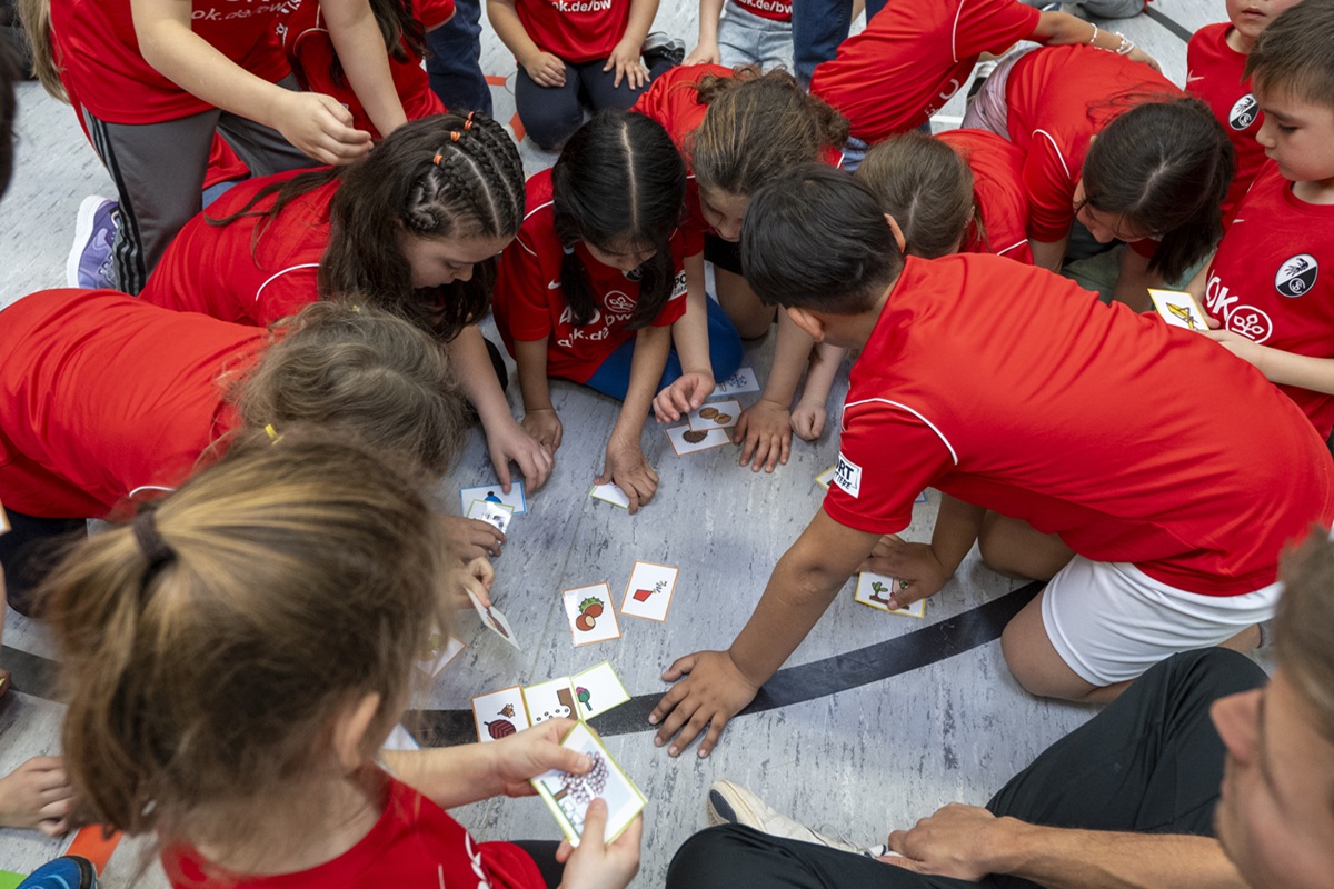 Viele Kinder in roten Trikots sitzen auf dem Hallenboden um Karten auf dem Boden.