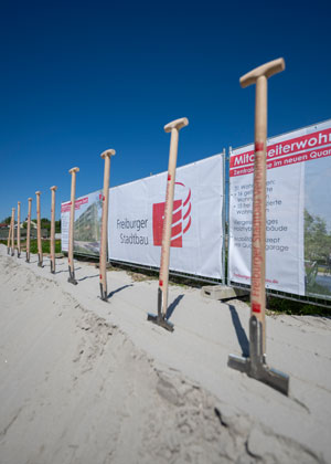 Mehrere Spaten stecken im Sand, im Hintergrund Bauzäune