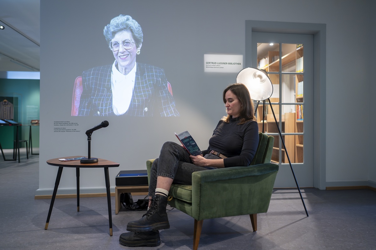 Eine junge Frau sitzt in einem grünen Sessel und liest eine Broschüre. Im Hintergrund ein Ausschnitt aus einem Video.