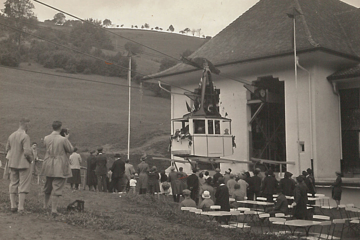 Schwarz-Weiß-Bild von der ersten Fahrt der Schauinslandbahn. Eine Gondel, auf der Wiese davor stehen und sitzen Menschen, die die Fahrt beobachten. 