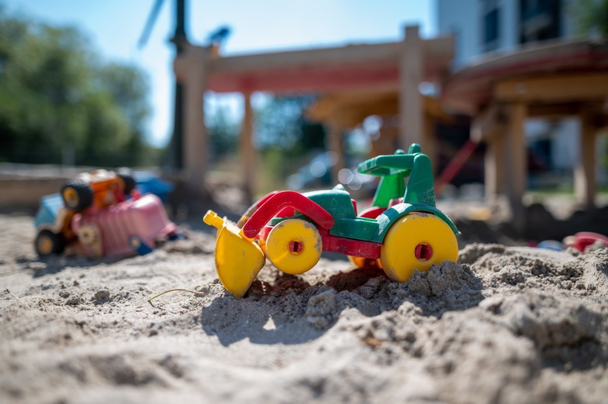 Nahaufnahme eines Spielzeug-Baggers im Sandkasten, im Hintergrund ein Spielplatz
