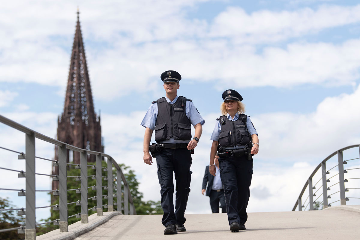 Zwei Mitarbeitende des Vollzugsdienstes auf einer Brücke, im Hintergrund das Münster.