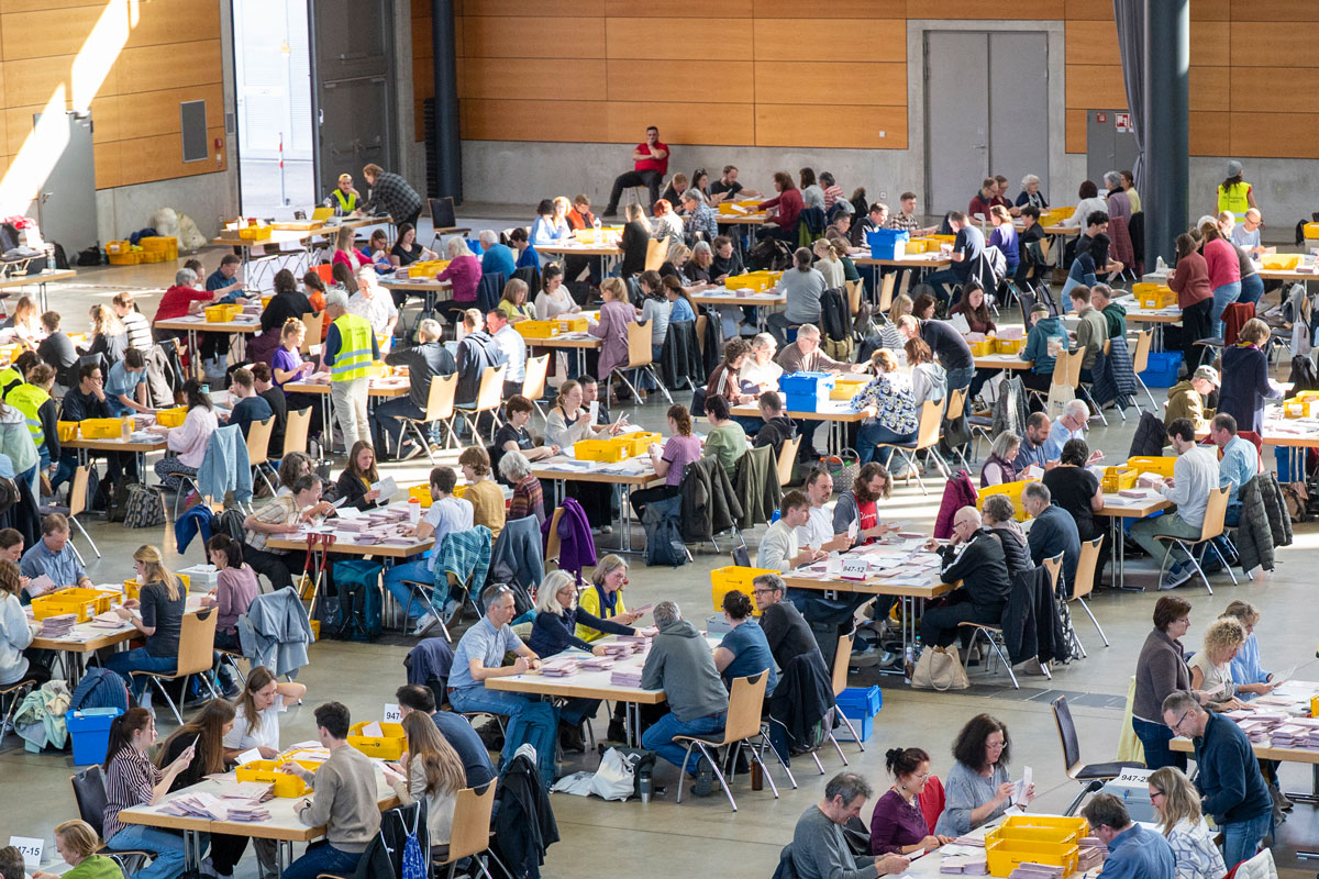 Viele Menschen sitzen in einer großen Halle an Tischen und zählen Wähler*innen-Stimmen aus