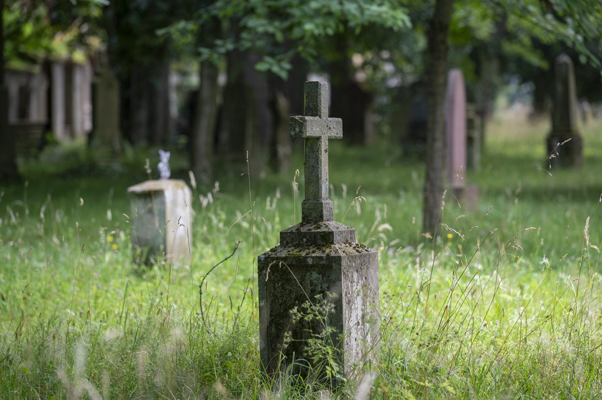 Eine grüne Wiese mit Bäumen, in der Mitte und im Hintergrund stehen zwei Grabsteine, der eine mit einem Kreuz darauf.