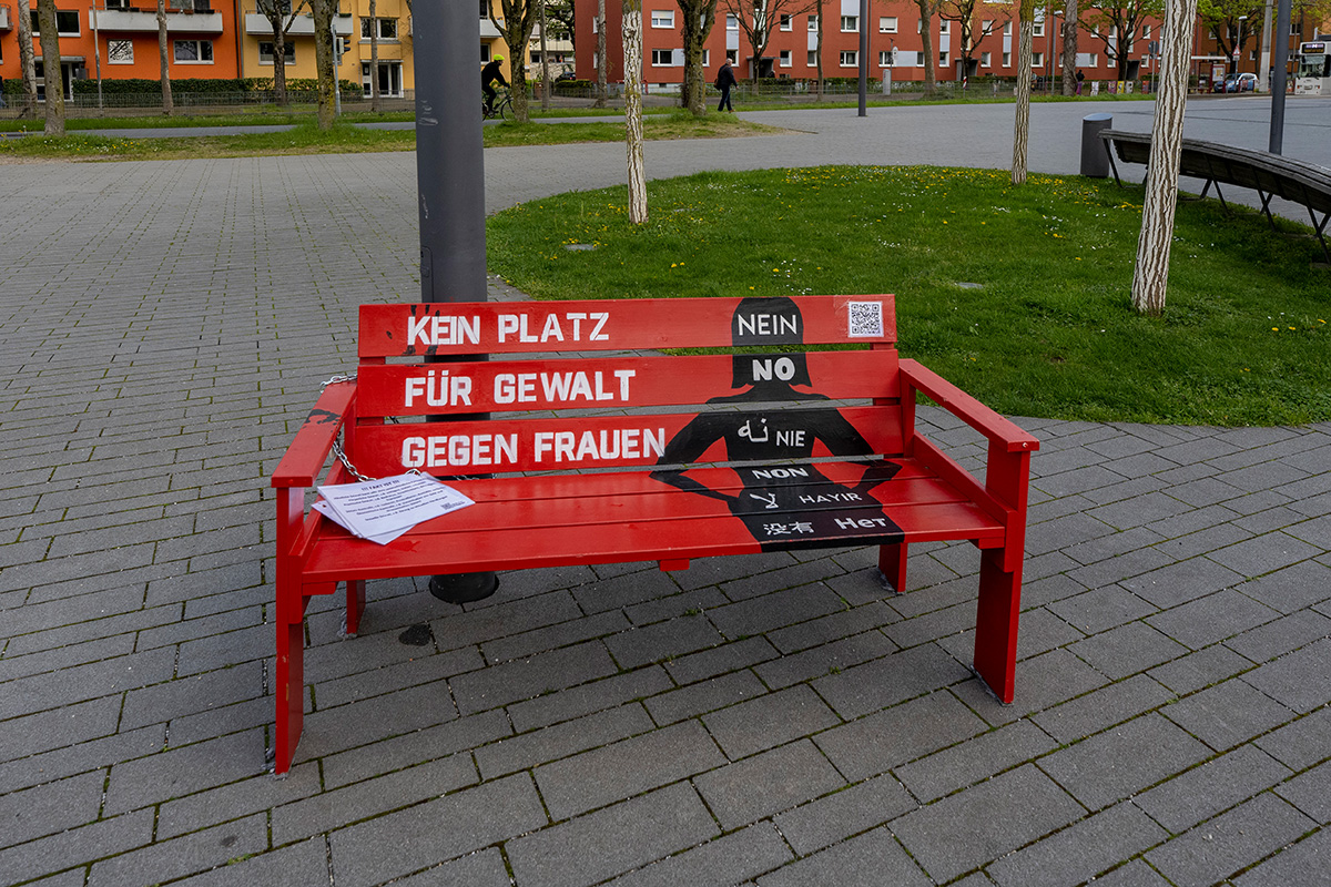 Rote Bank auf Pflastersteinen, darauf zu sehen ist der Text "Kein Platz für Gewalt gegen Gewalt an Frauen". dazu eine aufgemalte Silhouette einer Frau, au der in verschiedenen Sprachen das Wort "Nein" steht. 
