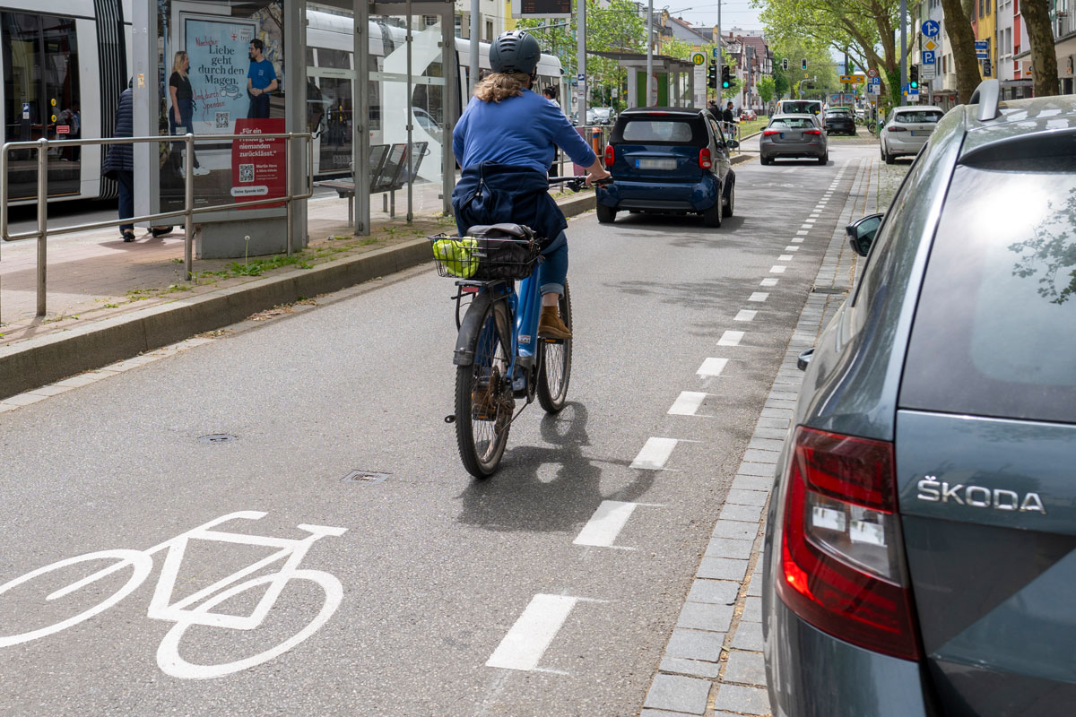 Person fährt mit Fahrrad, daneben parkende Autos und ein gestrichelter Streifen dazwischen