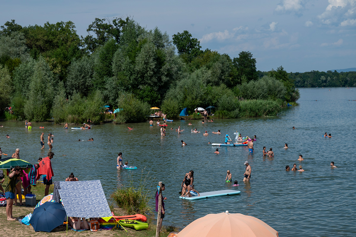 See mit Badenden im und am Wasser, am Ufer Sträucher und Bäume