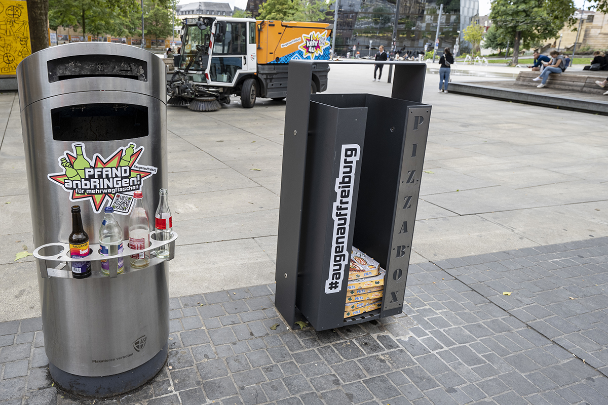 Runder Mülleimer mit Pfandring in dem Flaschen abgestellt werden können und eckiger Mülleimer mit Pizzakartons in der Innenstadt Freiburgs.