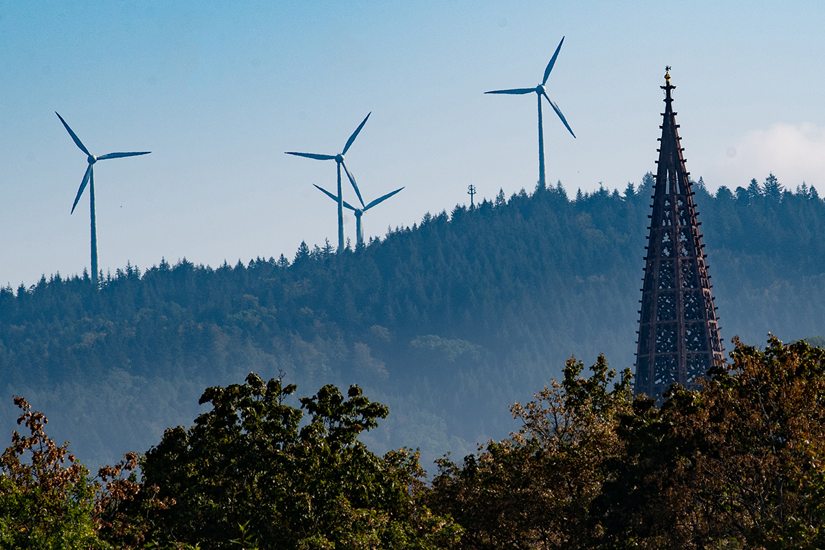 Vier Windräder stehen in der Ferne auf einem Berg, im Vordergrund Bäume und die Münsterspitze