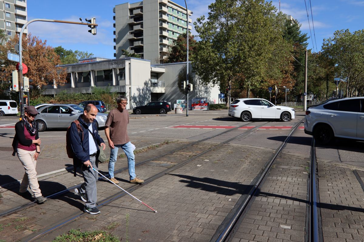 Drei Personen mit Blinden-Gehstock überqueren Bahnschienen auf der Fehrenbachallee