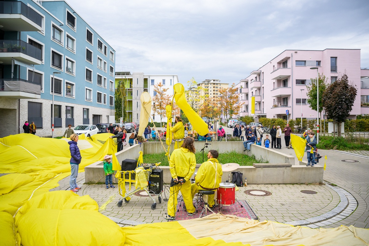 Innenhof im Quartier Gutleutmatten. Menschen in gelben Ganzkörperanzügen machen Kunst. Einer sitzt am Schlagzeug, ein anderer steht mit einem gelben Einkaufswagn daneben. Ein dritter steht in der Mitte mit drei gelben Keschern in der Hand. Viele Zuschauer*innen schauen zu. Der Boden ist mit einer gelben Folie bedeckt.