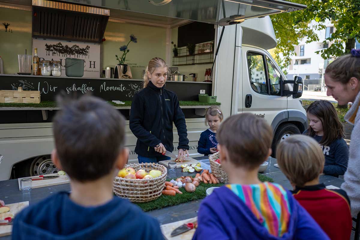 Kinder schauen Frau beim Gemüseschneiden zu, im Hintergrund Foodtruck