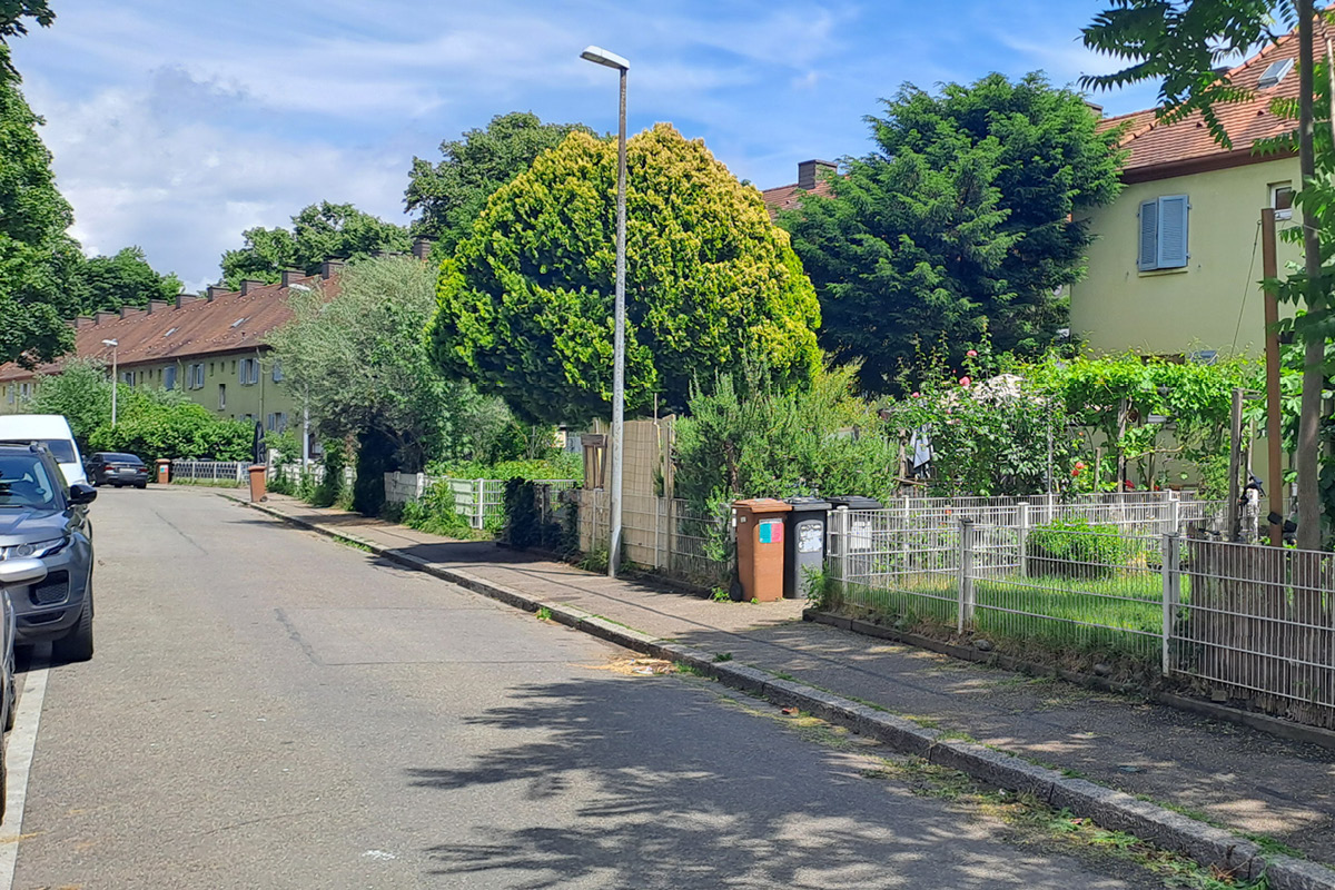 Gebäude und Gärten entlang der Carl-Mez-Straße (Bereich 2) Blick entlang der Carl-Mez-Straße im Bereich 2. Entlang der Straße stehen niedrige, zweigeschossige Wohnhäuser mit geneigten Ziegeldächern. Vorgärten mit Bäumen, Sträuchern und Blumen sind durch Metallzäune vom Gehweg abgegrenzt. Am Straßenrand stehen geparkte Autos und Mülltonnen; eine Straßenlaterne ragt über den Gehweg.