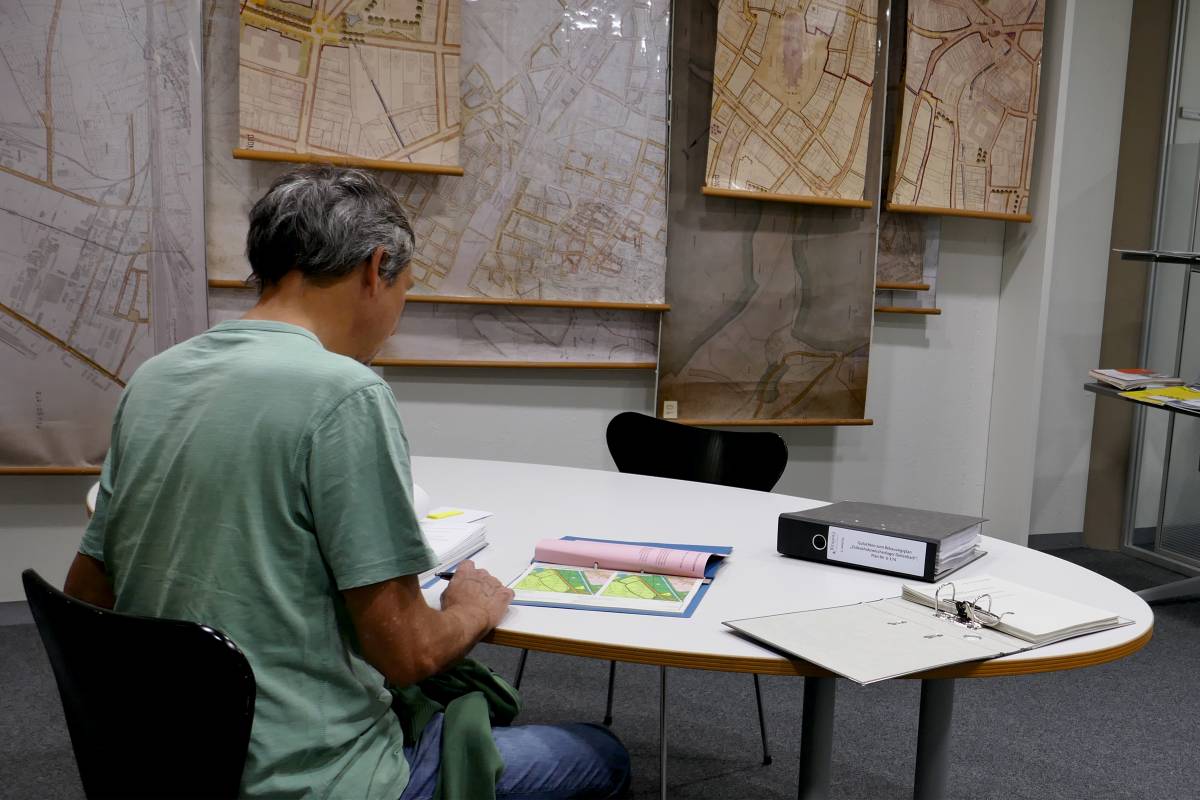 Mensch schaut sich am Tisch Pläne und Unterlagen an. An der Wand hängen viele Pläne