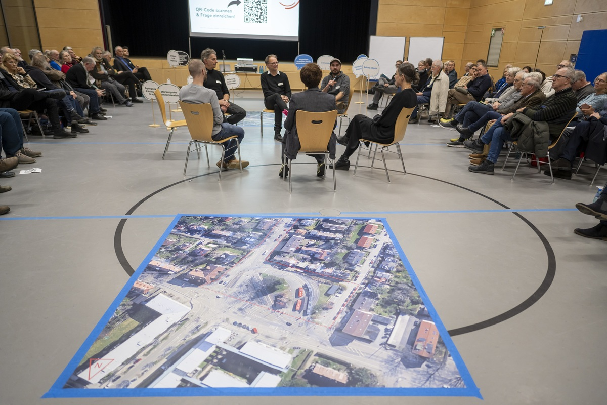 Sechs Personen unterhalten sich im Stuhlkreis. Ein großes Luftbild befindet sich auf dem Fußboden. Links und rechts sitzen Zuschauergruppen