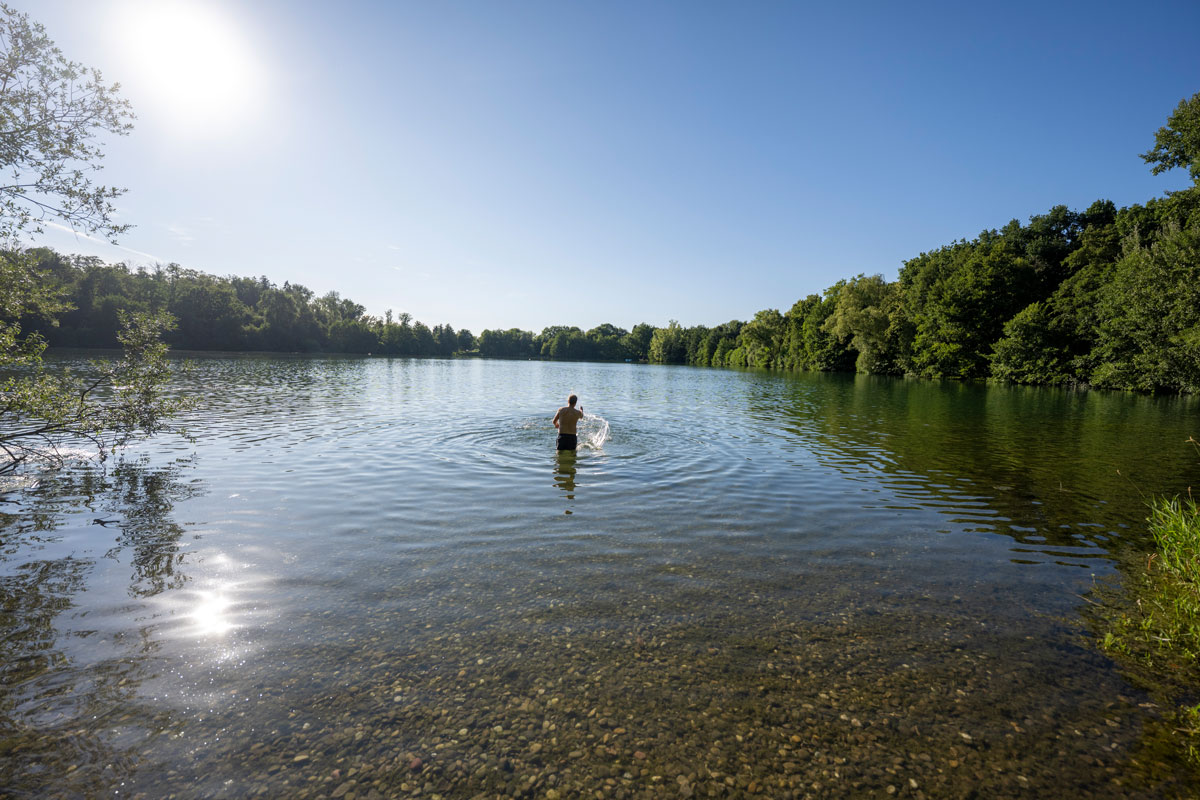 Person steht in Ufernähe im Wasser eines Badesees