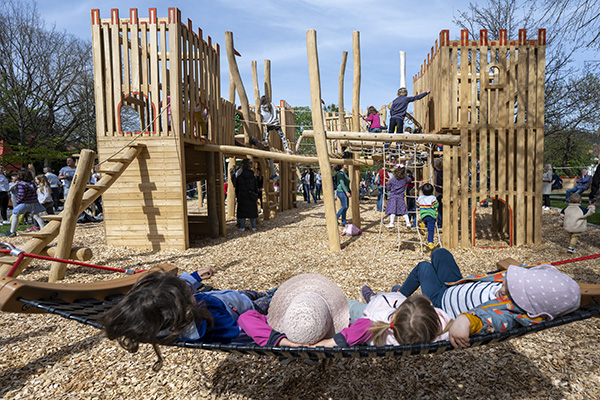 Kinder liegen auf einer Schaukel, im Hintergrund ein Klettergerüst und viele spielende Kinder