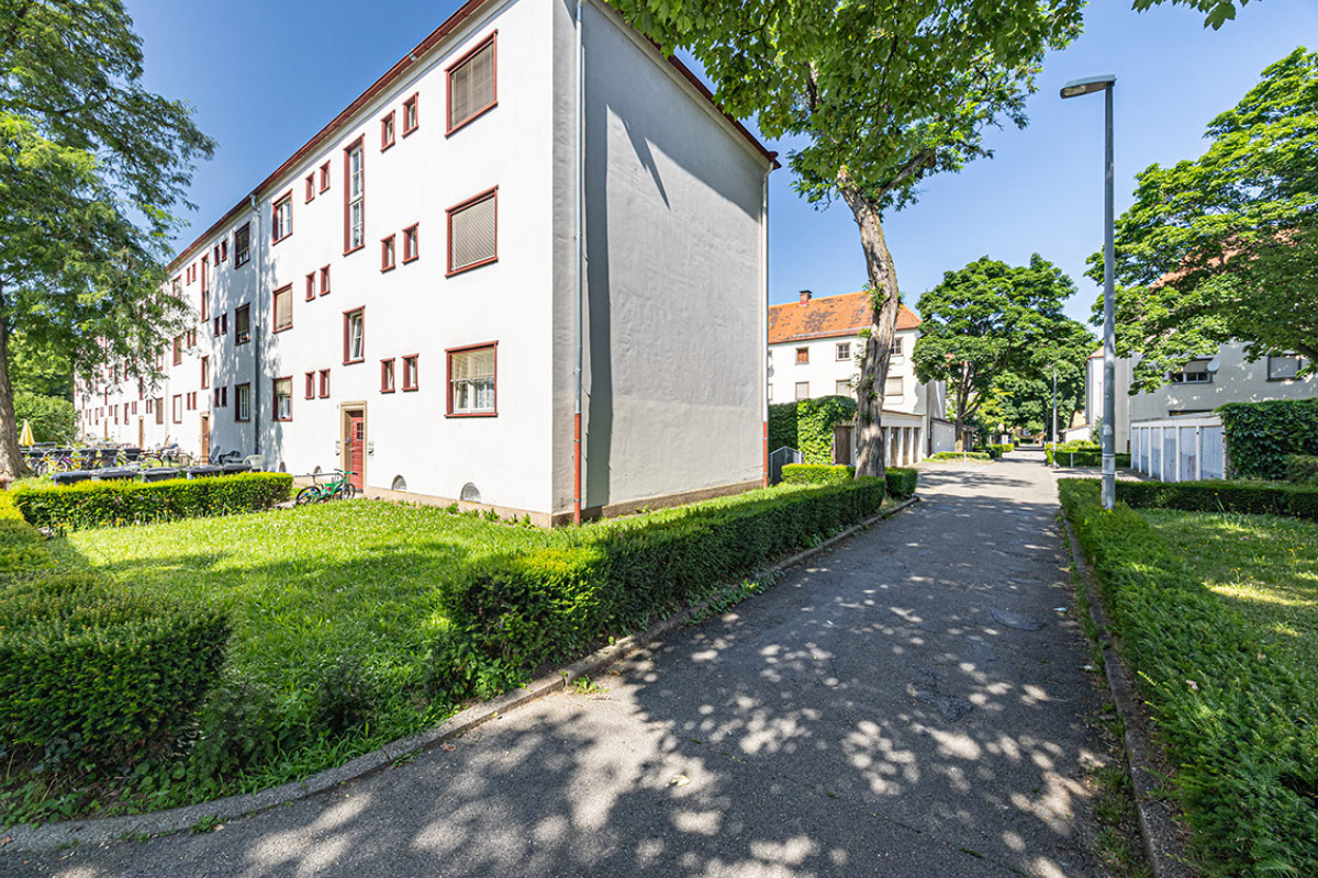 Fußweg zwischen denkmalgeschützten Gebäuden (Bereich 1) Ein asphaltierter Weg führt zwischen Grünflächen und niedrigen Hecken hindurch in ein Wohnquartier. Links steht ein hell verputztes, denkmalgeschütztes dreigeschossiges Wohnhaus mit rotbraun umrahmten Fenstern; rechts säumen Bäume, Rasen und Garagen den Weg. Eine Straßenlaterne steht am Wegesrand, im Hintergrund sind weitere Wohnhäuser zu sehen