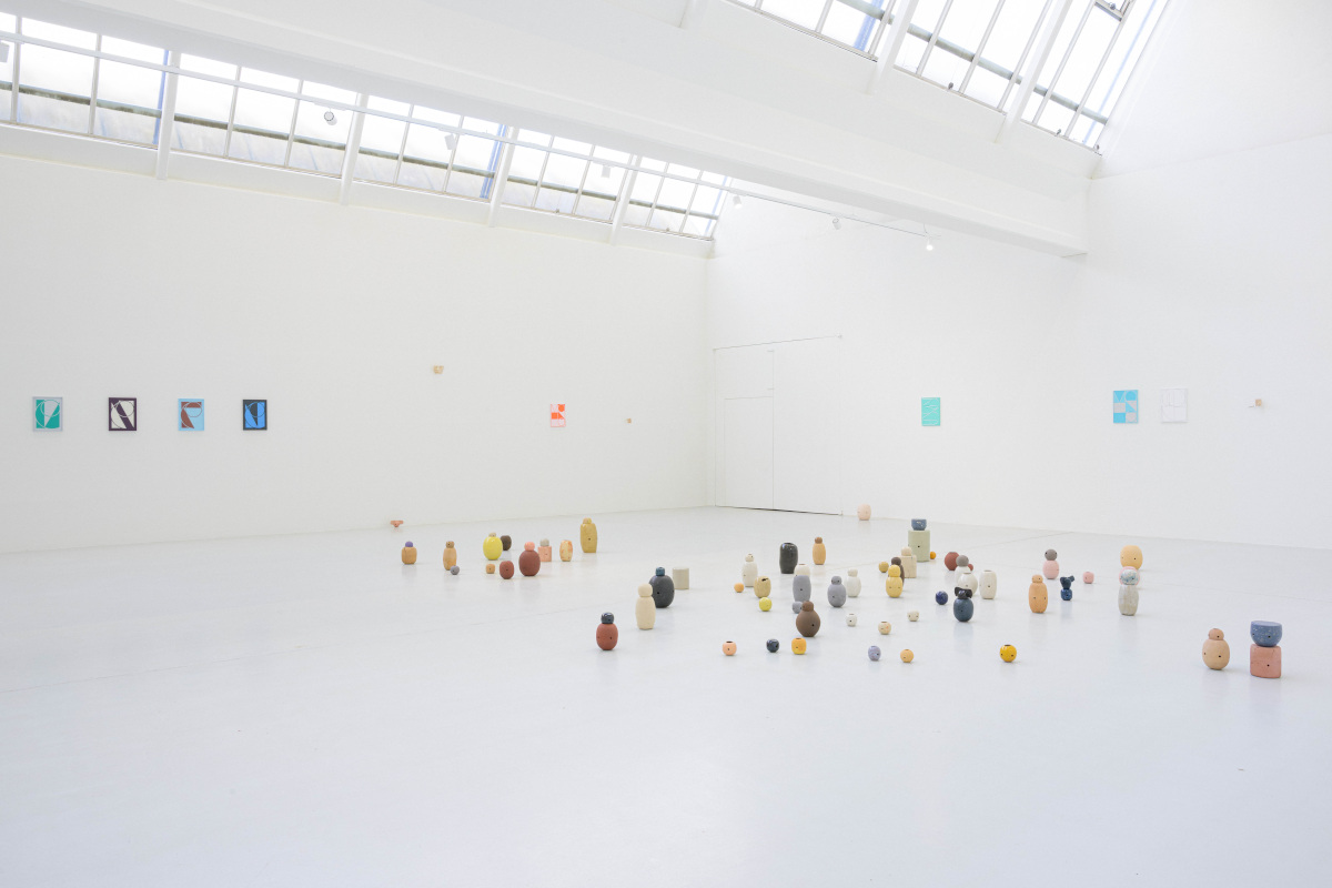 Ausstellungsansicht von "Following – some kind of same – π". Auf dem hellen Galerieboden sind zahlreiche kleine keramische Skulpturen in organischen Formen und gedeckten Farben (Ocker, Grau, Rosa, Gelb) frei verteilt. An den weißen Wänden hängen kleinformatige Gemälde mit grafischen Motiven.
