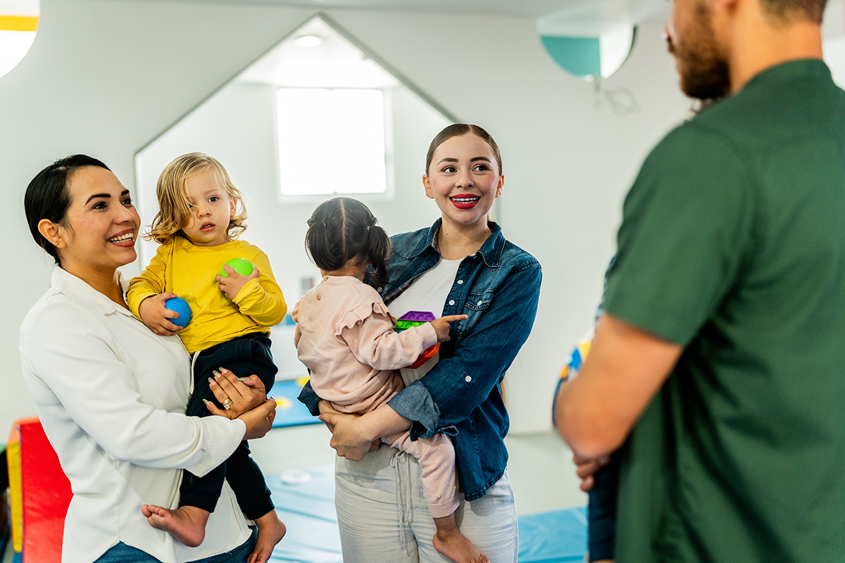 Zwei Mütter und ein Vater haben jeweils ein Kind auf dem Arm und unterhalten sich