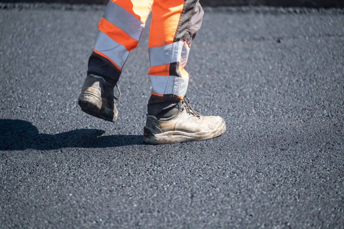 Unterschenkel eines Bauarbeiters mit Schutzhose und Arbeitsschuhen, steht auf einer geteerten Straße.