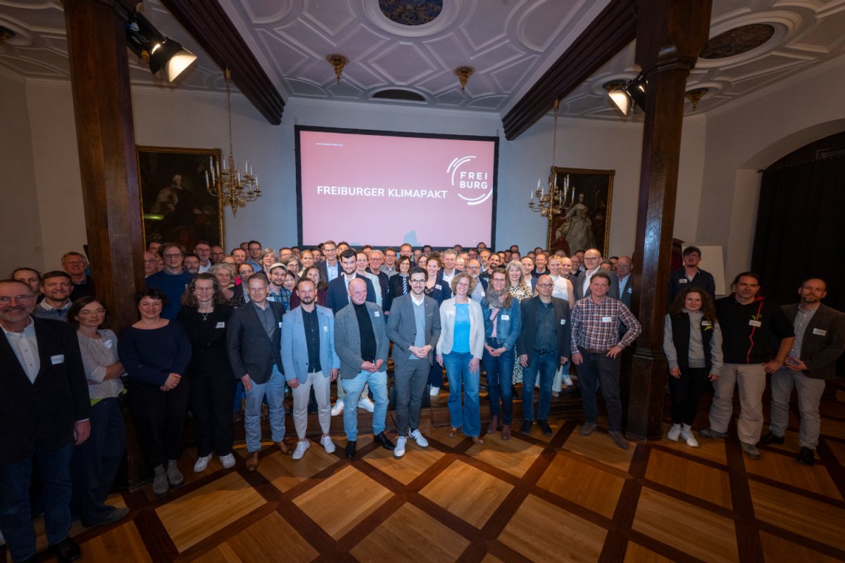 Eine große Ansammlung von Menschen steht in einem Saal im Halbkreis um eine Kamera, aus deren Perspektive das Foto gemacht wurde Im Hintergrund ist durch einen Beamer im der Schriftzug "Freiburger Klimapakt" projiziert