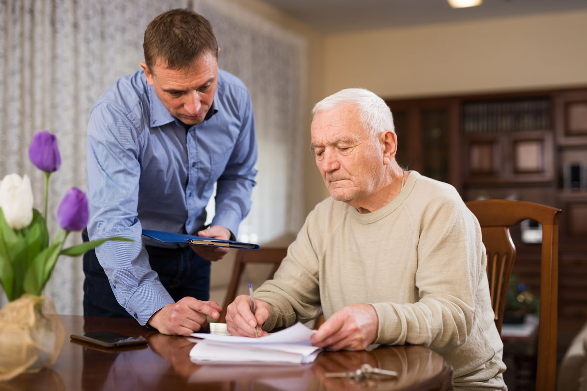 Ein Mann sitzt am Tisch und füllt etwas aus, ein anderer Mann steht neben ihm und hilft ihm dabei