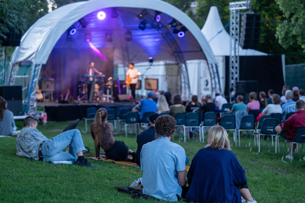 Festival: Bühne mit Musiker_innen, davor sitzt Publikum im Gras