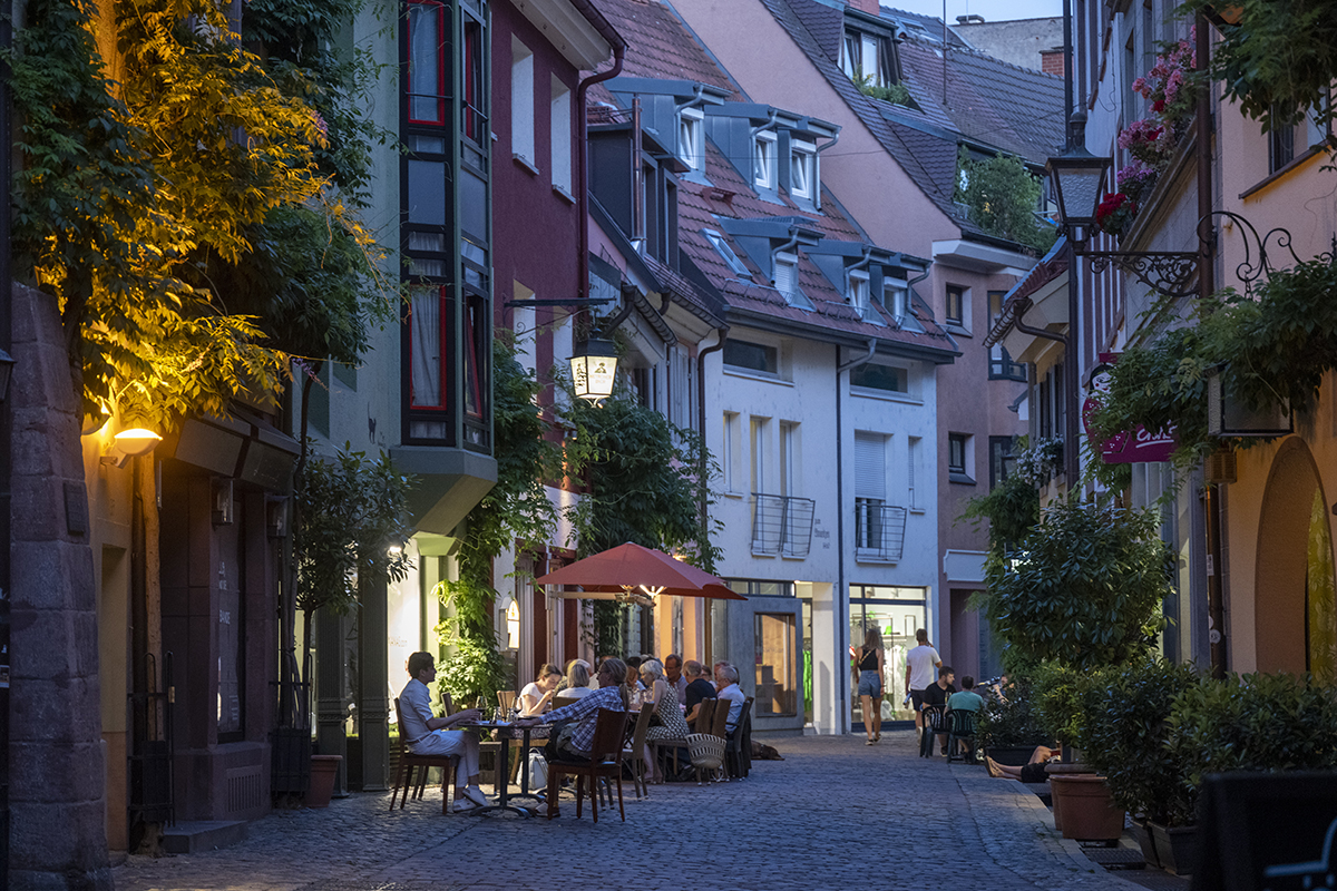 Außengastronomie in einer Seitenstraße Freiburgs; Menschen sitzen an Tischen