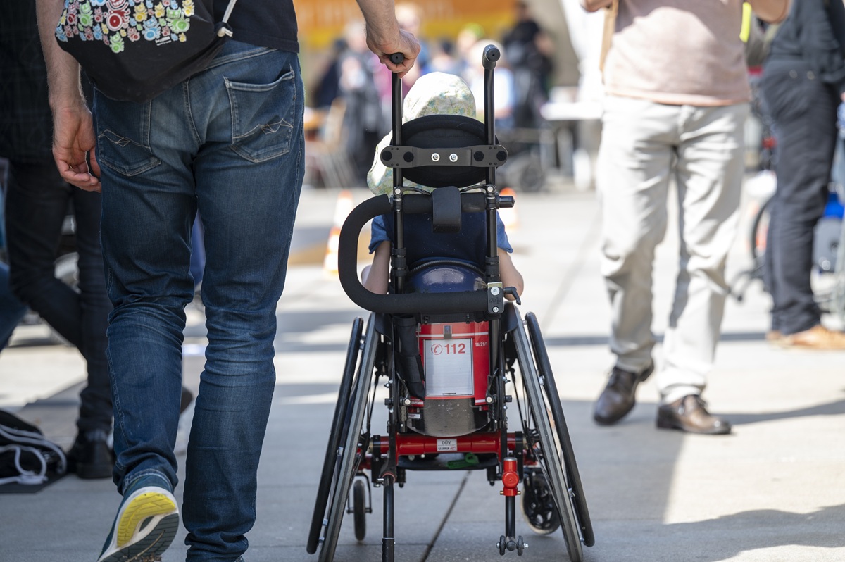 Das Bild zeigt ein Kind im Rollstuhl von hinten, das von einer erwachsenen Person geschoben wird.