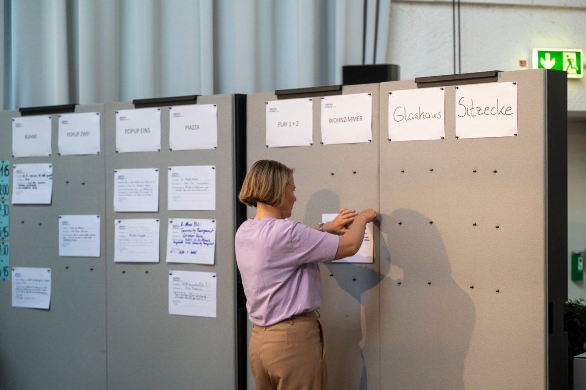 Eine Frau pinnt einen Zettel an eine graue Wand mit weiteren Zetteln. In der ersten Reihe oben stehen die Treffpunkte, in den Zeilen darunter mögliche Besprechungsthemen.