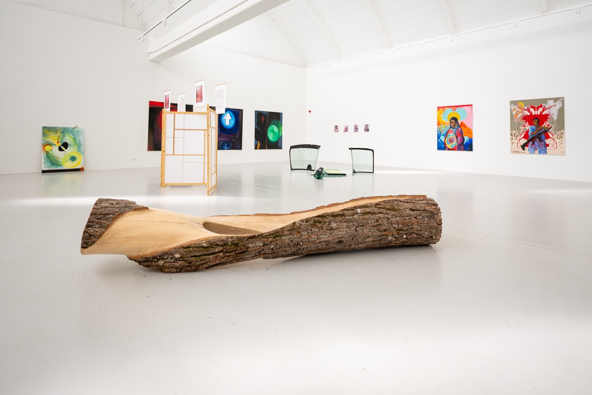 Ausstellungsansicht mit verschiedenen Kunstwerken: Ein großer bearbeiteter Pappelstamm zum Teil mit Rinde, im Hintergrund Malereien und eine Installation mit Autoglasscheiben