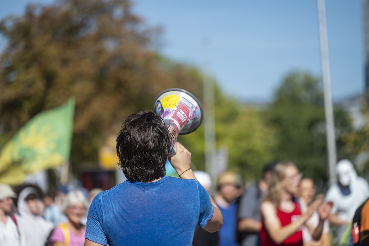 Eine FridayforFuture Demo, im Vordergrund eine Person mit blauem T-Shirt und einem Megafon in der Hand.