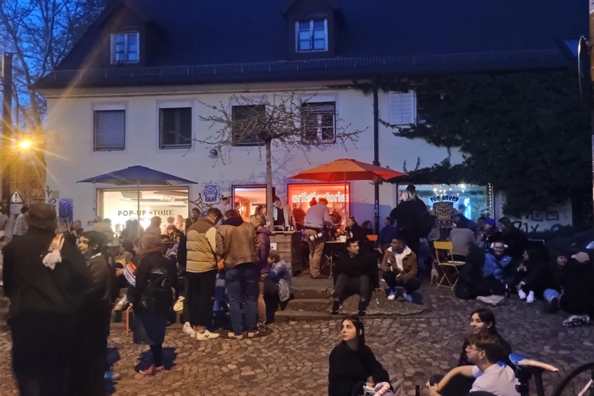 Pop-Up Store am Schwabentor, Foto: FWTM, Thorsten Schäfers Abendstimmung - Menschen stehen an Stehtischen auf einem Platz vor einem Haus mit beleuchteten Fenstern eines Ladenlokals